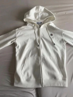 Vit moncler huvtröja - Snygg vit hoodie från Moncler med dragkedja framtill, huva med dragsko och två fickor. Klassisk Moncler-logga på bröstet. Tillverkad i mjukt bomullsmaterial som känns skönt mot huden. Perfekt för en clean och stilren look.