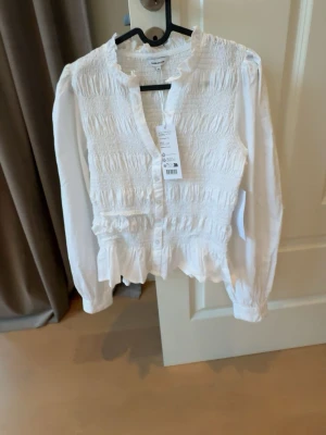 Vit smockad blus med volang - Säljer denna supersöt vit blus med smockad överdel, volangkant vid hals och nederkant samt knappar! Aldrig använd, fick i present! Säljer den då för att jag inte riktigt tyckte att den passade mig. Storlek Xs! Tyvärr som de syns på sista bilden är de en liten sminkfläck från när jag har provat den❣️