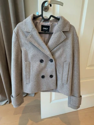 Beige/grå kappa med svarta knappar - Säljer en kappa från ONLY då den inte kommer till använd för mig, storlek S! Den är knappt använd! Den har svarta knappar, och en knapp på insidan av ena sidan för att stänga till lite extra!