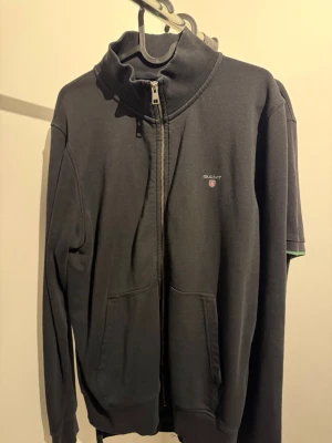 Svart zip hoodie från GANT - Svart hoodie från GANT med dragkedja framtill och två fickor. Klassisk design med ribbade muddar och hög krage. Liten GANT-logga med sköld på bröstet. Tillverkad i mjukt bomullsmaterial för en skön känsla.