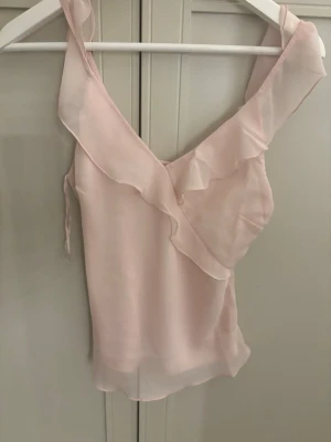 Ljusrosa volang linnetopp - Supersöt ljusrosa linnetopp med volangdetaljer och v-ringning. Tunt, luftigt material som ger en romantisk känsla. Perfekt för varma dagar och lätta outfits. Axelbanden är smala och toppen har en feminin siluett.