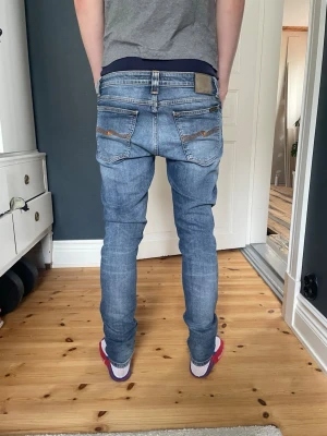 Blå nudie jeans - Säljer ett par klassiska blå jeans med normal passform och snygga slitningar. Jeansen har fem fickor, knappgylf och detaljerade sömmar på bakfickorna. Perfekta för en avslappnad stil och tillverkade i bomull.