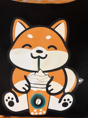 Svart hoodie med söt shiba-tryck - Svart hoodie med stor, färgglad shiba inu-illustration på bröstet där hunden dricker bubble tea. Tryck i orange, vitt och svart. Ficka framtill och huva med dragsko. Orange text och kaffe-motiv på ärmen. Perfekt för dig som gillar gulliga och unika plagg.