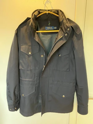  Polo Ralph Lauren field jacket  - Snygg mörkblå field jacket  från Polo Ralph Lauren med fyra framfickor, dragkedja och tryckknappar. Jackan har hög krage och justerbara ärmslut. Tillverkad i slitstarkt material som passar perfekt för blåsiga dagar.