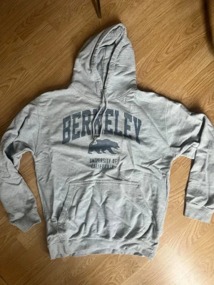 Grå Berkeley hoodie med tryck - Snygg grå hoodie med stor huva och dragsko. På bröstet står det 'BERKELEY' i mörkblått med en björn och texten 'University of California' under. Klassisk känguruficka framtill och ribbade muddar vid ärmar och midja. Perfekt för chill dagar eller plugget. Är storlek M men sitter mer som S