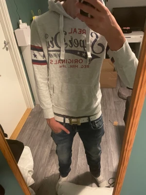 Grå Superdry hoodie med tryck - Säljer en grå hoodie från Superdry med stort tryck på bröstet och röda samt blå detaljer. Hoodien har huva med dragsko, känguruficka och ribbade muddar. Perfekt för dig som gillar streetwear och vill ha en skön och snygg tröja.