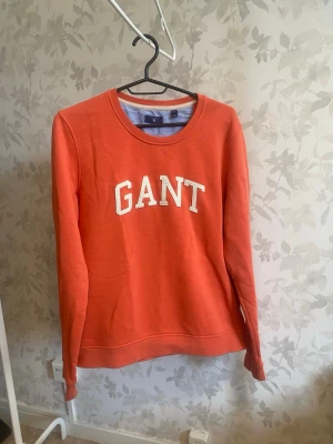 Orange sweatshirt från GANT - Snygg orange sweatshirt från GANT med klassisk rund halsringning och vit broderad logga på bröstet. Tröjan har långa ärmar och är gjord i mjukt bomullsmaterial. Perfekt för en avslappnad och trendig look.