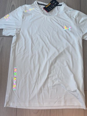 Vit t-shirt från Under Armour - Snygg vit t-shirt från Under Armour med holografiska loggor och detaljer på bröstet, ärmen och längs sidan. Tillverkad i ett lätt och mjukt material som passar perfekt till träning eller chill. Klassisk rund hals och korta ärmar.