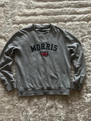 Morris sweatshirt med flagga i grå - stl M - Morris sweatshirt i gråmelerat, storlek M. 100% Bomull, rund hals med ribbade muddar och nederkant, broderad Morris-text och Union Jack på bröstet.  Använd men i mycket fint skick, inga hål eller fläckar. Personen på bilden är 189cm, väger 78kg.  Rök- och djurfritt hem. Skriv gärna om du vill ha fler bilder eller undrar något!