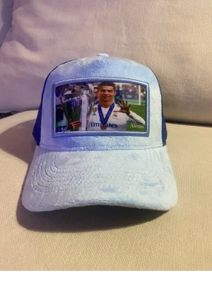 Blå Ronaldo keps - Säljer en ljusblå och mörkblå truckerkeps med mesh på sidorna och baksidan. Framtill finns ett fototryck av Ronaldo..