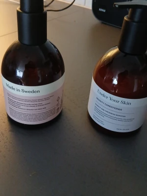 Under Your Skin Density Conditioner - Säljer en brun plastflaska med pump, innehållande Under Your Skin Density Conditioner. Balsamet är tillverkat i Sverige och kommer i en stilren, minimalistisk design med vit etikett. Perfekt för dig som vill boosta hårets volym och tjocklek.