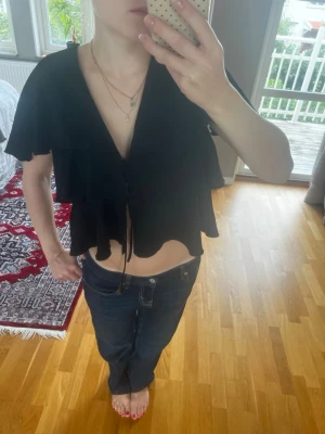 Svart volangblus med knyt - Säljer en svart blus med flera volanglager och knyt framtill. Blusen har korta ärmar och en lös passform som ger en flowy känsla. Perfekt för dig som gillar en romantisk och trendig look.