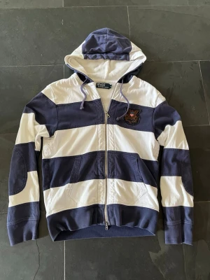 Vintage polo Ralph Lauren hoodie - Säljer en riktigt fet vintage Polo Ralph Lauren hoodie i storlek M. Den har riktigt schysst passform och har coola detaljer såsom koppar broderiet på bröstet. Är gjord i Sydkorea under tidigt 2000 tal. Tröjan har mindre defekter i ärmsluten och någon fläck på framsidan. Hör gärna av dig för mer bilder eller andra funderingar! 100% bomull