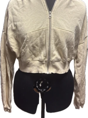 Beige croppad hoodie med dragkedja - Säljer en beige croppad hoodie med dragkedja framtill och två stora fickor. Hoodien har långa ärmar, ribbade muddar och en avslappnad passform. Perfekt för en chill och trendig look. Materialet är mjukt och skönt bomullstyg.