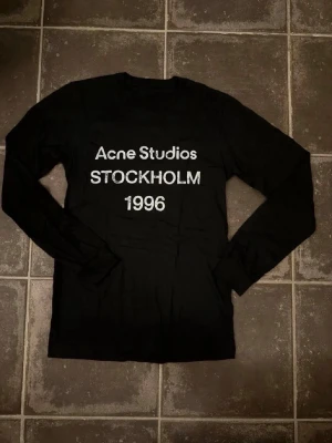 Svart långärmad Acne Studios t-shirt - Svart långärmad t-shirt från Acne Studios med vit text 'Acne Studios STOCKHOLM 1996' på bröstet. Klassisk rund halsringning och rak passform. Perfekt för dig som gillar stilrena och trendiga plagg. Acne är lite oversize men den passar som en vanlig M