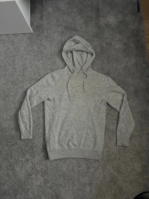 Ljusgrå merinoull hoddie - En stilren ljusgrå hoodie med huva och dragsnören. Tillverkad i mjukt, stickat material som känns skönt mot huden. Slim passform och passar personer från 160-173 skulle jag säga.  Skick enligt bilderna