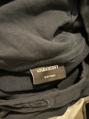 Svart hoodie från Lyle & Scott - Säljer en svart hoodie från Lyle & Scott med klassisk gul logga broderad på ärmen. Hoodien har ribbade muddar och känguruficka framtill. Perfekt för dig som gillar stilrena och bekväma plagg. Materialet är mjukt och skönt mot huden.