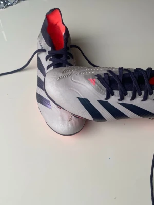 Adidas Predator fotbollsskor silver - Snygga Adidas Predator fotbollsskor i silver med svarta och lila detaljer samt röda inslag på dobbar och insida. Skorna har snörning och är tillverkade i syntetmaterial med texten 'HYBRIDTOUCH 2.0' på sidan. Perfekta för dig som vill sticka ut på planen. Storleken på skorna är 40 och ett halvt och är pros. Helt ny skick har aldrig använt de ute. Råka beställa för liten storlek därför säljer jag skorna 