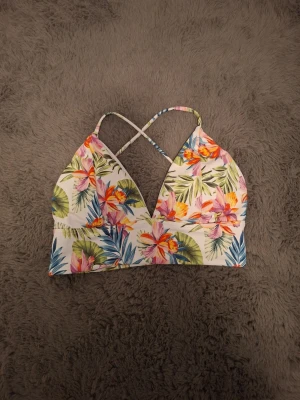Blommig bikiniöverdel från Stadium - Säljer en färgglad bikiniöverdel från Stadium i storlek 38. Överdelen har tropiskt blommönster i rosa, orange, grönt och blått på vit botten samt korsade axelband i ryggen. Materialet är mjukt och stretchigt, perfekt för strand eller pool.