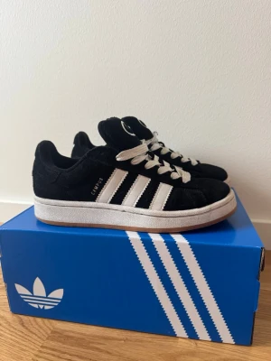 Adidas Campus 00 svarta  - Sparsamt använda, säljes eftersom jag fått större fötter och inte längre passar storlek 38. Ett par klassiska Adidas Campus 00 sneakers i svart mocka med vita linjer. Skorna har vita snören som är lite smutsiga. Medföljer även svarta oanvända snören. Perfekta för en clean och stilren look.