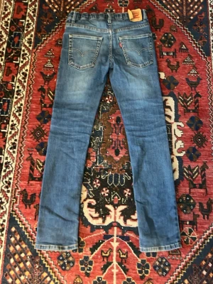 Blå raka jeans från Levi's - Säljer ett par klassiska blå Levi's jeans med raka ben och femficksmodell. Jeansen har tydliga sömmar på bakfickorna och en ikonisk läderpatch i midjan. Tillverkade i slitstark denim med en snygg tvättad look. Perfekta för en avslappnad och tidlös stil.