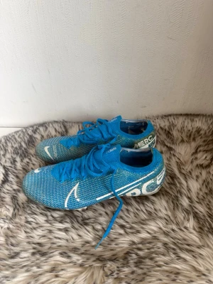 Blå Nike Mercurial fotbollsskor - Säljer ett par blå Nike Mercurial fotbollsskor med vita detaljer och texten 'JUST DO IT' på sidan. Skorna har snörning och en låg profil, samt mönstrad ovandel i syntetmaterial för bra bollkontroll. Perfekta för dig som vill sticka ut på planen.