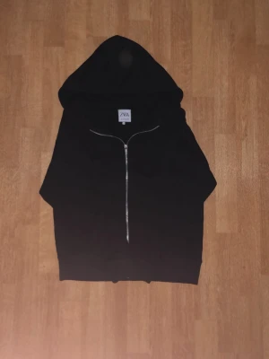 Svart hoodie med dragkedja från ZARA - En svart hoodie från ZARA med hel dragkedja framtill och stor huva. Klassisk och enkel design utan tryck eller mönster, perfekt för en avslappnad stil. Tillverkad i mjukt material som känns skönt mot huden.
