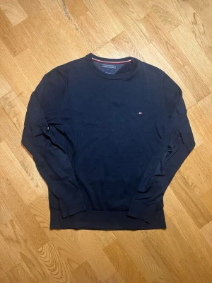 Mörkblå stickad tröja Tommy Hilfiger - Säljer denna svarta stickad tröja från Tommy Hilfiger med rund halsringning och diskret logga på bröstet. Tröjan har långa ärmar och ribbade muddar vid ärmslut och nederkant. Perfekt till en avslappnad men stilren look. Vid frågor, extra bilder eller liknande är det bara att kontakta mig!