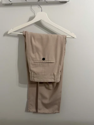 Beige chinos med bakficka - Snygga beige chinos med klassisk rak passform och en bakficka med knapp. Byxorna har hällor för bälte och är tillverkade i ett mjukt, vävt material som ger en stilren look. Perfekta för dig som gillar en clean och enkel stil.