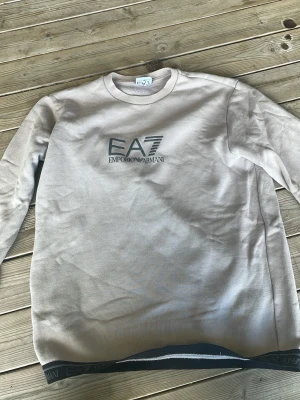 Beige sweatshirt från Emporio Armani - Snygg beige sweatshirt från Emporio Armani EA7 med tryckt logga på bröstet och dekorativt band med logotyp längs nederkanten. Tröjan har rund halsringning och är tillverkad i mjukt material, perfekt för en avslappnad och trendig look.
