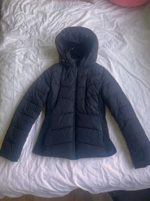 Svart figursydd Zara jacka - Stilren svart pufferjacka med stor huva och dragkedja framtill. Jackan har en figurnära passform, quiltade sömmar och två sidofickor. Perfekt inför hösten och vintern. Den ger en modern look med sin matta finish.