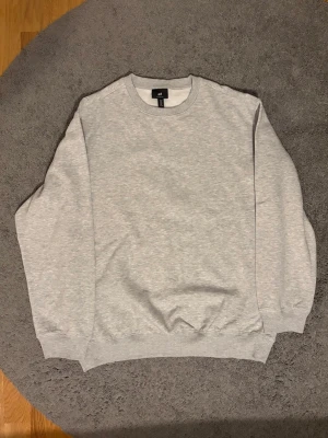 Ljusgrå sweatshirt från H&M - En basic ljusgrå sweatshirt från H&M med rund halsringning och ribbade muddar vid ärmslut och nederkant. Perfekt för en avslappnad och enkel stil. Tillverkad i mjukt material som känns skönt mot huden. Lite oversized så sitter lite mer som L