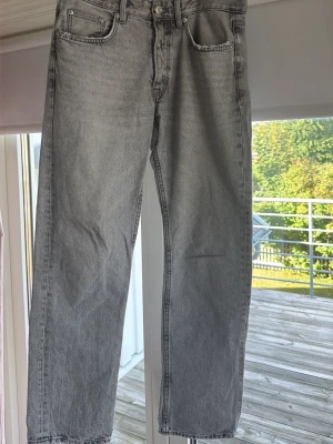 Grå raka jeans med rå kant - Säljer ett par grå jeans med rak passform och rå kant längst ner. Jeansen har klassisk femficksdesign och knappgylf. Perfekta för en avslappnad och trendig look.