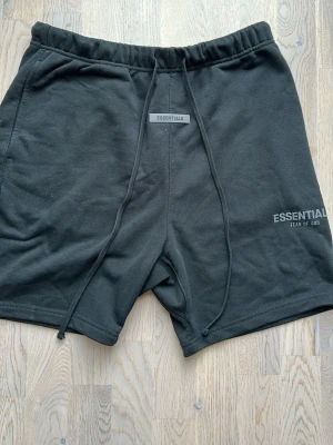 Svarta shorts Essentials Fear of God - Snygga svarta shorts från Essentials Fear of God med dragsko i midjan och logga tryckt på vänster ben. Tillverkade i mjukt bomullsmaterial för en bekväm känsla. Perfekta för sommaren och har en avslappnad passform.