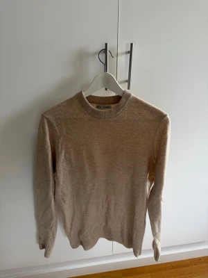 Beige stickad tröja från Zara - Snygg beige stickad tröja från Zara med rund halsringning och ribbade muddar. Tröjan har en enkel och stilren design som passar perfekt till många olika outfits. Materialet ser mjukt och bekvämt ut, perfekt för kyligare dagar.