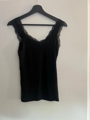 Svart linnetopp med spetsdetaljer - Snygg svart linnetopp med v-ringning och spetsdetaljer längs axelbanden och ringningen. Toppen är ärmlös och har en feminin, stilren look som passar till många olika outfits. Perfekt att styla lager på lager eller bära som den är.