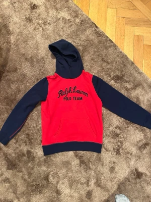 Röd och marinblå hoodie Polo Ralph Lauren - Snygg hoodie från Polo Ralph Lauren med röd kropp och marinblå ärmar och huva. Framsidan har broderad text 'Ralph Lauren Polo Team'. Klassisk känguruficka och ribbade muddar. Perfekt för en avslappnad och sportig stil.