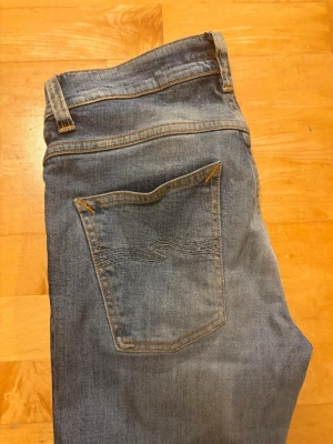 Nudie - Säljer ett par klassiska blå jeans med slim passform och en snygg vågig söm på bakfickan. Jeansen har normal midja och är gjorda i slitstarkt denimtyg. Perfekta för en avslappnad och tidlös stil. Modellen är grim Tim, jeansen är uppsydda till L28.