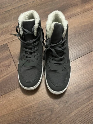 Gråa vinterkängor med foder - Säljer ett par svarta höga kängor med vit sula och mjukt vitt foder. Kängorna har snörning framtill och en sportig look, perfekta för kalla dagar. Insidan är fodrad med syntetpäls för extra värme och komfort.