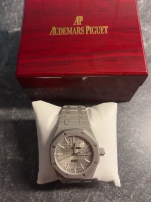 Audemars Piguet Royal Oak klocka silver - Helt ny och oandvänd AUTOMATISK Audemars Piguet Royal Oak klocka i silver med ikonisk åttakantig boett och länkarmband i metall. Urtavlan har ett rutigt mönster och silverfärgade visare och index. Kommer i trälåda med guldfärgad logga. INTE RIKTI
