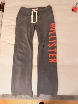 Gråa mjukisbyxor från Hollister - Säljer ett par gråa mjukisbyxor från Hollister med breda ben och vit dragsko i midjan. Byxorna har stora fickor och en stor röd logga längs ena benet. Perfekta för chill dagar hemma eller till träningen.