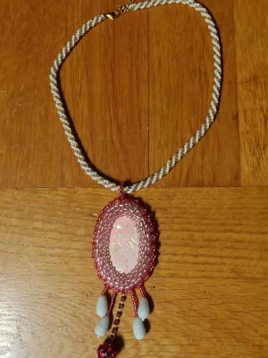 Handgjort halsband med pärlor och hänge - Unikt halsband med tvinnad kedja i vitt och beige. Stort ovalt hänge med rosa och genomskinliga pärlor runt en dekor med rosa och guld mönster. Nedanför hänget hänger vita droppformade pärlor och en rad rosa glittrande pärlor. Perfekt för dig som gillar statement-smycken.