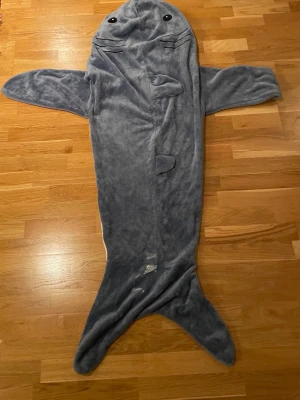 Hajdräkt i fleece för maskerad - Rolig hajdräkt i mjuk grå och vit fleece med fenor, stjärt och huva med ögon och gälar. Perfekt för maskerad eller cosplay, dräkten täcker hela kroppen och har detaljer som gör att du ser ut som en haj.