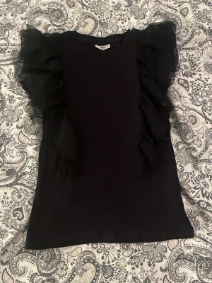 Svart topp med volangärmar - Snygg svart topp med stora volangdetaljer längs ärmarna som ger en cool och trendig look. Toppen är ärmlös med volanger i tunt tyg som sticker ut och gör outfiten extra unik.💗