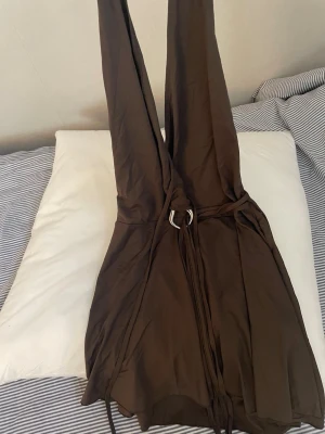 Brun omlottklänning med djup ringning - Snygg brun omlottklänning med djup v-ringning och knytband i midjan. Klänningen är ärmlös och har en kort längd, perfekt för varma dagar. Tillverkad i ett mjukt och följsamt material som ger en skön passform.