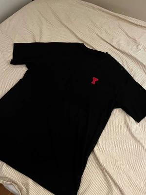 Svart Ami paris t-shirt - Snygg svart t-shirt med ett litet rött broderat hjärta på bröstet. Klassisk passform och rund halsringning. Perfekt basplagg som funkar till det mesta och är enkel att matcha med andra plagg. Ami Paris.