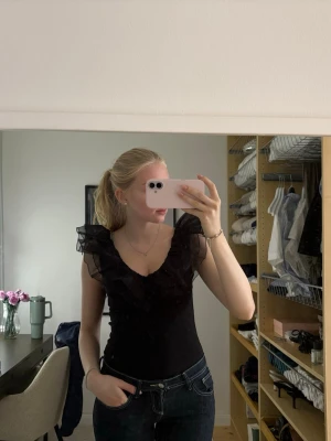 Svart body med volang  - Snygg svart body med djup v-ringning och volangdetaljer i mesh över axlarna. Toppen är ärmlös och har öppen rygg, vilket ger en trendig och feminin look. Perfekt att styla med jeans eller kjol för en cool outfit.