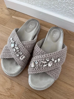 Beige sandaler med pärlor från XIT - Snygga beige sandaler från XIT med breda, virkade band i kors och glittrande pärldekorationer. Mjuka innersulor och öppen tå ger en avslappnad men trendig look. Perfekta för dig som vill ha något extra till sommaren.