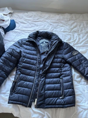 Svart dunjacka från Superdry - Snygg svart dunjacka från Superdry, modell Fuji Jacket. Jackan har quiltad design, hög krage och två dragkedjeförsedda fickor framtill, ena är trasig. Perfekt för kalla vinterdagar och har en modern, sportig look.