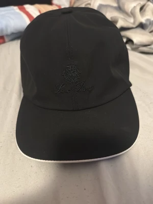 Svart keps Loro piana - Stilren svart keps från Loro piana med broderad logga och text framtill. Kepsen har böjd skärm med vit kant och elastiskt band bak för skön passform. Materialet ser ut att vara mjukt och lätt, perfekt för att lyfta din outfit.